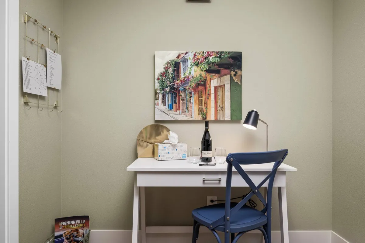 A calm, well-lit workspace with a desk, chair, desk lamp, and a bottle of wine, decorated with tasteful artwork. The setup suggests a peaceful place for focused work, journaling, or creative pursuits, illuminated by gentle natural light.