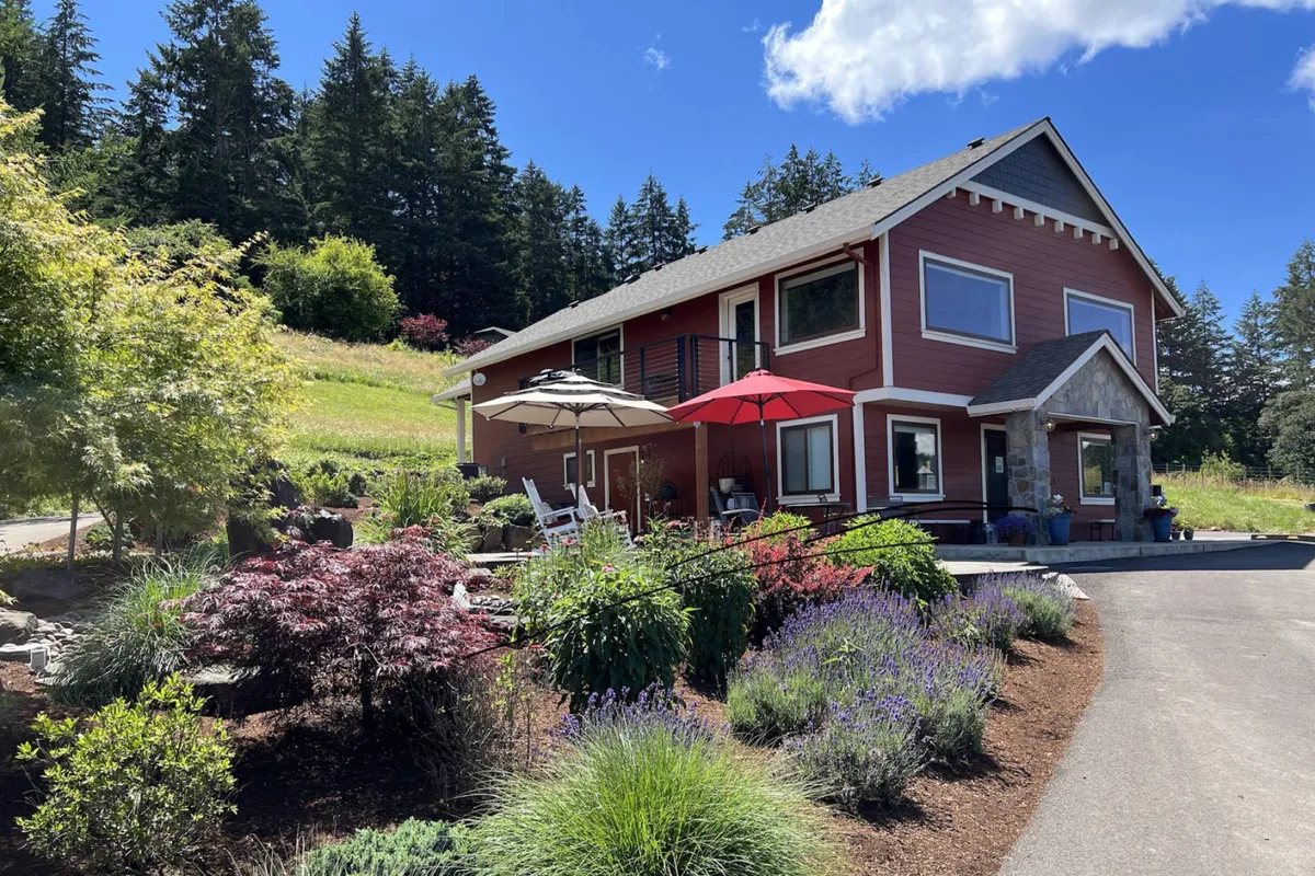 The image showcases a vibrant red farmhouse set against rolling fields, with well-tended gardens, inviting patio umbrellas, and a warm glow from the setting sun. The scene exudes tranquility and hospitality, inviting guests to relax outdoors and soak in the natural beauty of Oregon wine country.
