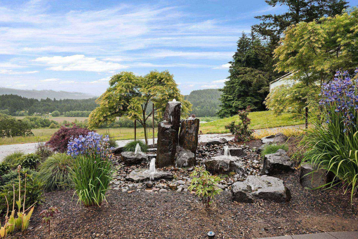 A peaceful setting featuring a rock fountain surrounded by colorful flowers and lush greenery. The scene is serene and celebrates the presence of nature and seasonal blooms.
