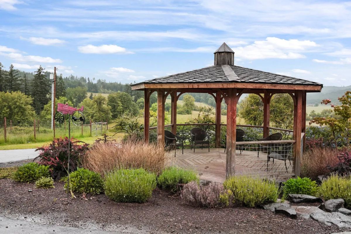 The image showcases an elegant gazebo set within a landscaped garden, surrounded by blooming flowers and soft hills. The serene atmosphere is ideal for relaxing, reading, or gathering with friends, capturing the essence of a peaceful country retreat.