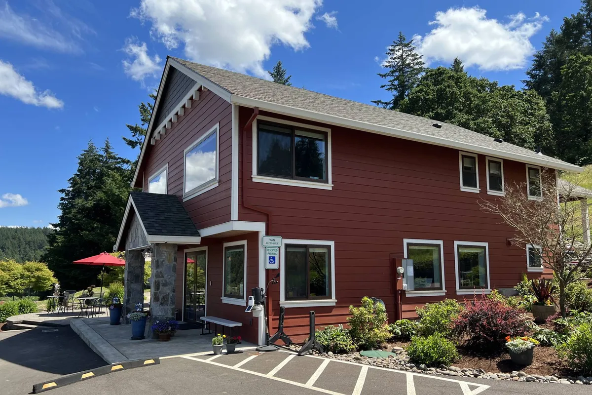 This image captures the welcoming red exterior of the farmhouse, accented by stone details and lush greenery. The patio area suggests a space for relaxing or socializing outdoors, surrounded by vibrant nature.