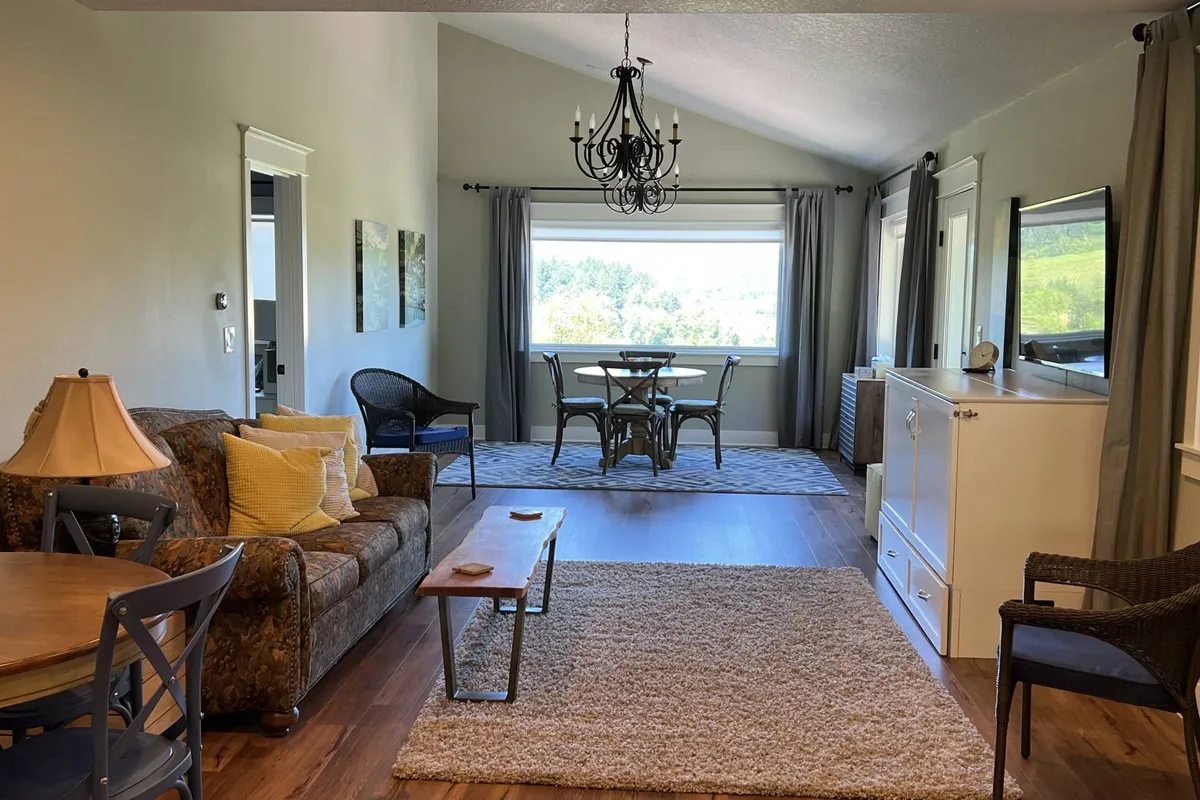 A charming space with a comfortable sofa, a dining table set near a large window, and a chandelier above, all bathed in natural light. The layout encourages both relaxation and socializing, while the window frames countryside views, blending comfort and scenery.