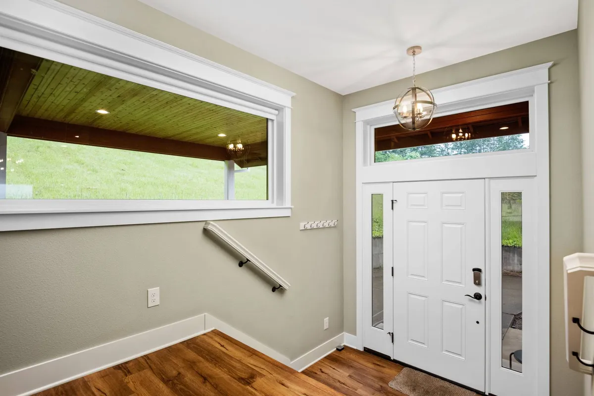 This image captures the home's welcoming entryway, featuring a front door bordered by a large window that lets in abundant natural light. The staircase in the background and the glimpse of the outdoors through the glass establish a strong sense of arrival and connection to the surrounding landscape.
