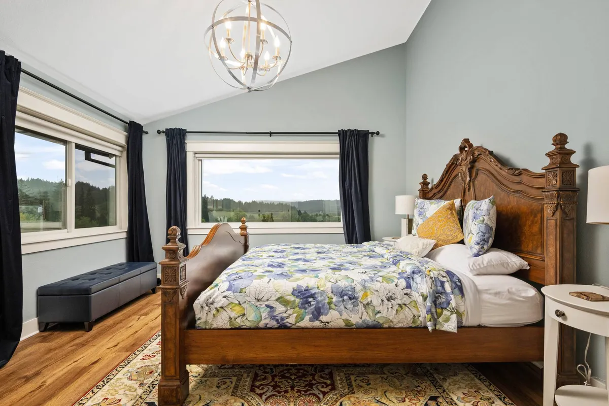 A serene bedroom space featuring an ornate wooden bed, decorative bedding, and large windows with picturesque views of rolling hills. The soft light and inviting atmosphere suggest a perfect spot to relax, read, or quietly reflect.