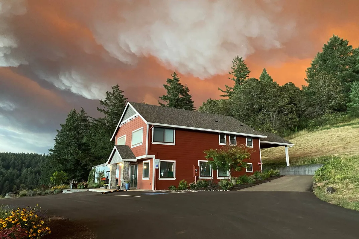 The exterior of the red house is nestled among green trees beneath a dramatic sky, with a peaceful driveway leading up to it. The porch offers vantage points for sunset watching and tranquil outdoor moments after a day of exploring.